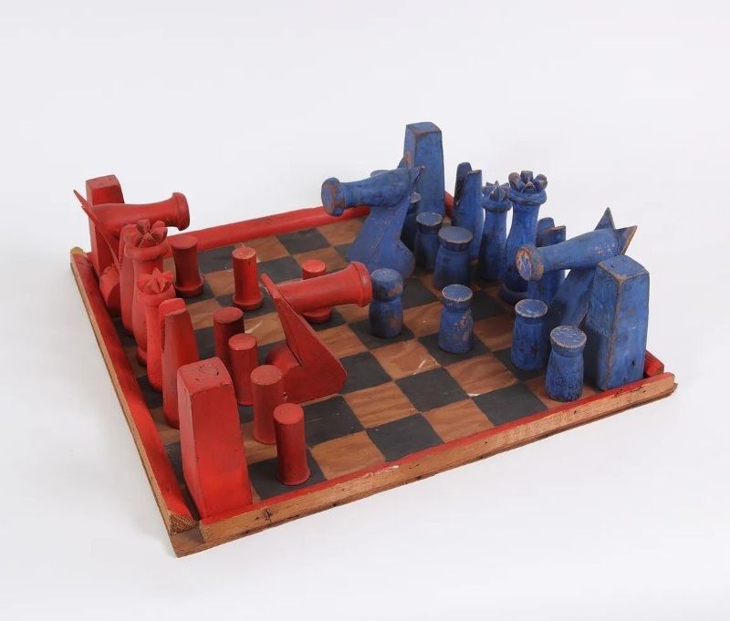 A wooden chess set with blocky red and blue pieces. The knights are about three times the size of the other pieces and dominate the board.