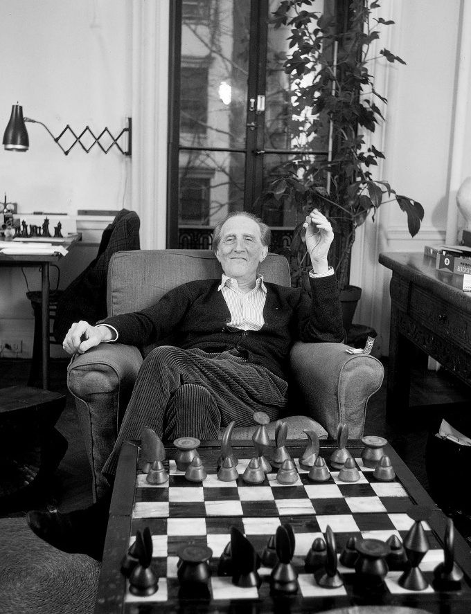 Marcel Duchamp lounges in an easy chair behind a modernist chess set, smiling at the camera and holding a cigar.