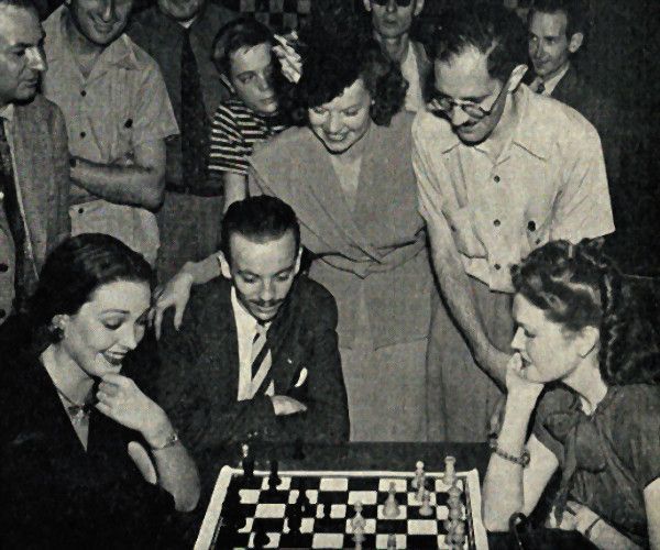 At the 1945 Pan American Chess Congress, Linda Darnell and Roseanne Murray play an off-hand game while Reuben Fine, Hector Rosetto, Mitzi Mayfair and Isaac Kashdan watch.