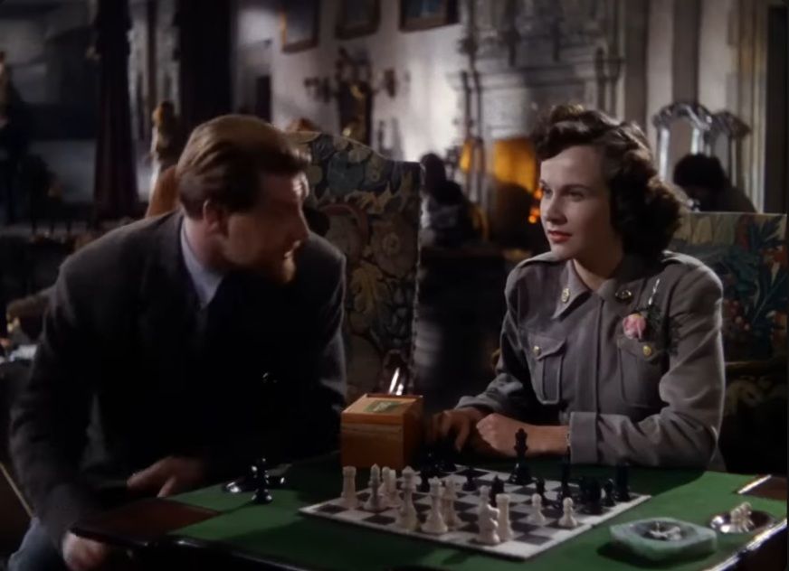 In a scene from A Matter of Life and Death, Kim Hunter sits at a green felted table and plays chess with a club-style plastic set. She wears a gray military uniform. Roger Livesey sits to the side of the table and leans in to speak to Hunter.