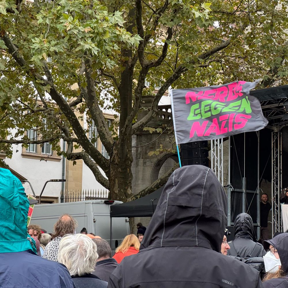 Demoteilnehmer:innen von hinten, vor der Bühne wird eine Flagge "NERDS GEGEN NAZIS" geschwenkt.