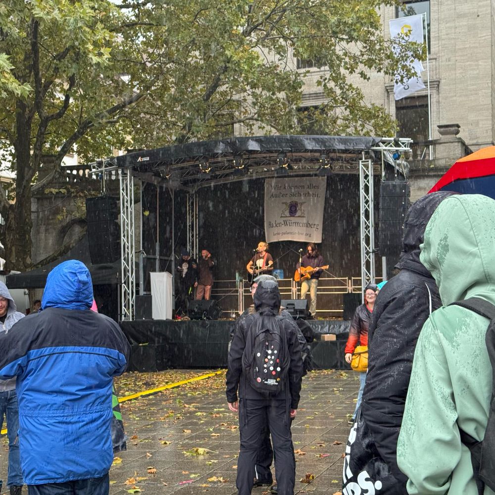 Die Musik Spiel auf der Bühne, aber davor regnet es in Strömen. Weniger Leute (die waren unter die Bäume geflohen), mehr Regenschirme. Im Hintergrund der Bühne hängt ein Banner: "Wir können alles. Außer Sicherheit."