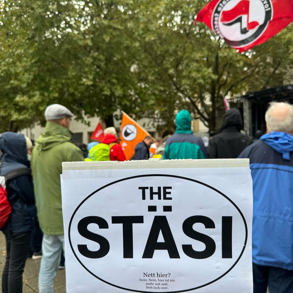 Demoschild "THE STÄSI" in Anlehnung an die Marketingcampagne "THE LÄND", im Hintergrung Demoteilnehmer:innen teils mit Flaggen (Linke, Piraten, Antifaschisten)