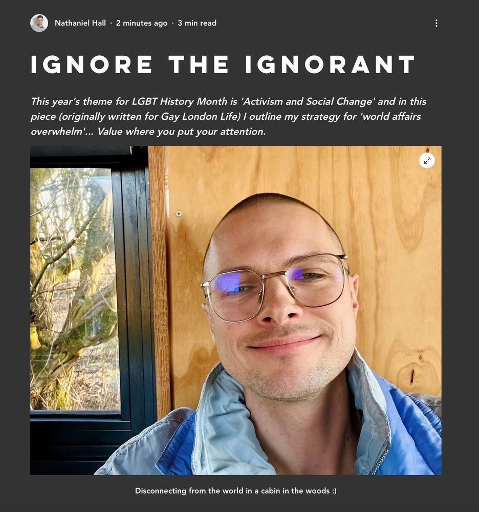 A photo of Nathaniel, a white gay man with shaved hair and glasses, sat in front of a wooden wall and window overlooking woodland. 