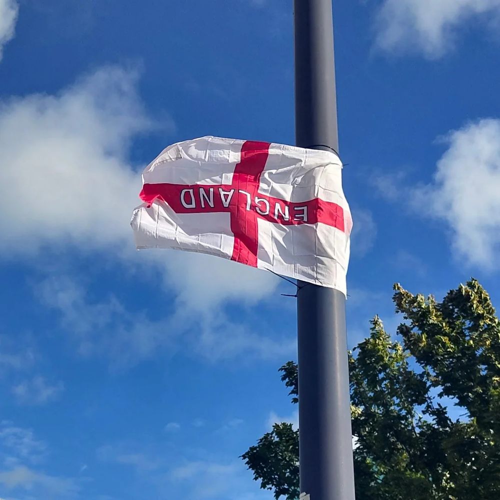 Photograph of the flag of England attached to a gray pole against a blue sky with white clouds. The flag is white with a red cross and the word 