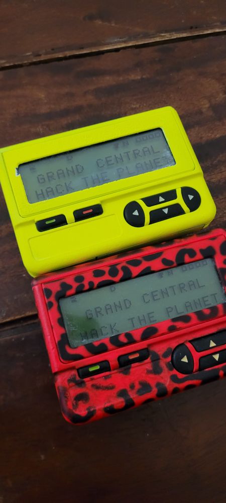 A neon yellow pager beside a leopard print neon pink pager, both displaying the message "GRAND CENTRAL HACK THE PLANET"