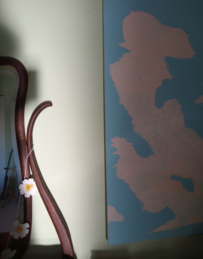 The edge of an old mirror, a painting of a cumulonimbus cloud. Felt daisies hang from the mirror. An electric bass guitar is on a stand reflected in the mirror. Shadows surround, except for a beam of late afternoon sunlight between the mirror and painting.
