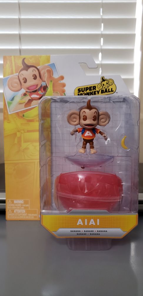 Figure of AiAi from Super Monkey Ball by Jakks Pacific in the package. He comes with a banana and a two piece ball he can roll around in, as well as a base for him to stand on while in the ball. The package is yellow and white, featuring artwork of AiAi as he's about to grab a banana and the Super Monkey Ball logo. The logo is specifically the same one used for Banana Mania, featuring AiAi's head.