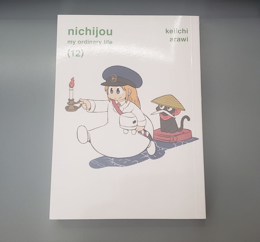 The cover of "Nichijou - Volume 12" is shown on a zoom up of a blue desk. Hakase is seen wearing a blue mailman hat and red mailbag, while holding a candle holder with her right hand and a plush of a white creature under her left arm. Sakamoto sits on top of her mailbag, wearing a bamboo hat and white mustache. Underneath them are shadows resembling various creatures.