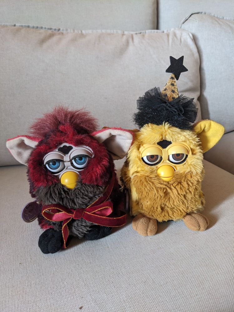 Twili furbies side by side. The one on the left is red and is wearing a velvet ribbon. The one on the right is yellow and wearing a glittery party hat.