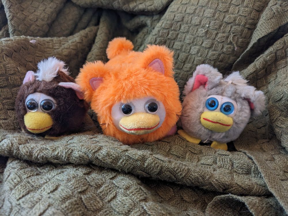 A photo of three toys. The left one is a brown Furby fake with grey eyes, the middle one is an orange cat peepy, and the right one is a grey Furby fake with blue eyes.