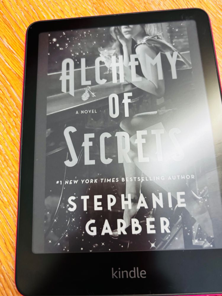  an e-reader (a Kindle) displaying the cover of a book titled “Alchemy of Secrets” by Stephanie Garber, who is noted as a #1 New York Times bestselling author.

The cover art features a woman sitting at what appears to be a bar, holding a cocktail glass, with sparkling or star-like effects around her — giving it a glamorous, possibly magical or mysterious vibe that fits the title.