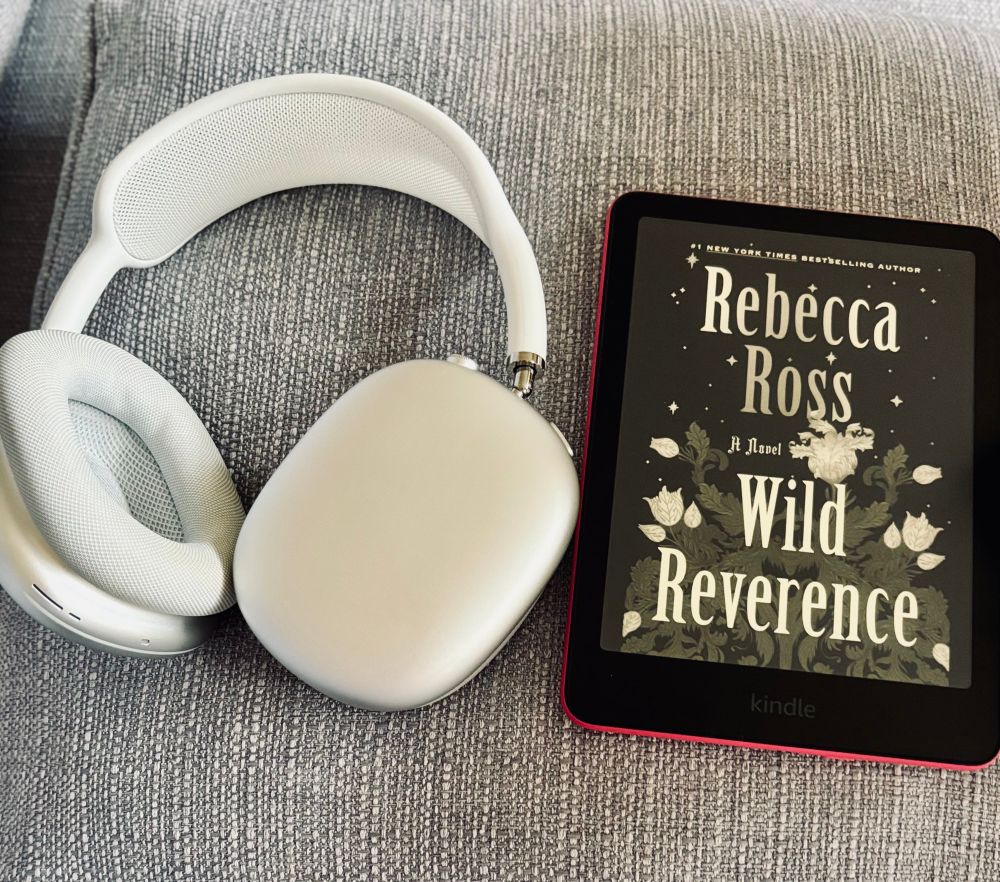 a pair of Apple AirPods Max headphones in silver/white placed on a gray fabric surface, next to a Kindle e-reader. The Kindle screen displays the cover of a book titled Wild Reverence by Rebecca Ross.
