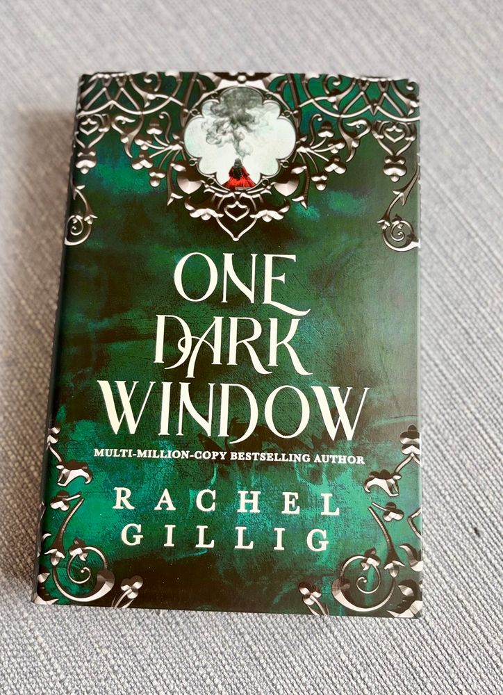 The photo shows the book One Dark Window by Rachel Gillig. The cover is dark green with ornate, silver decorative patterns around the edges and an illustration near the top showing a small figure in red with smoke rising above them. The text on the cover includes:
	•	Title: One Dark Window
	•	Author: Rachel Gillig
	•	Tagline: Multi-million-copy bestselling author
