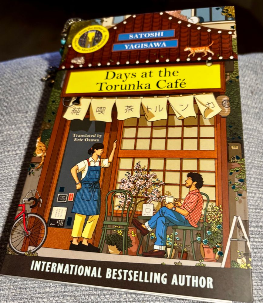 a book titled “Days at the Torunka Café” by Satoshi Yagisawa, translated by Eric Ozawa.
The cover illustration shows:
	•	A cozy café storefront with wooden sliding doors.
	•	A person in an apron standing at the entrance, as if greeting or talking to someone.
	•	Another person sitting outside at a table, drinking a hot beverage.
	•	Plants, a bicycle, and decorative elements around the café.
	•	A small illustrated cat on the roof sign.
	•	A banner with Japanese characters hanging above the door.

At the bottom, it also says “INTERNATIONAL BESTSELLING AUTHOR.”