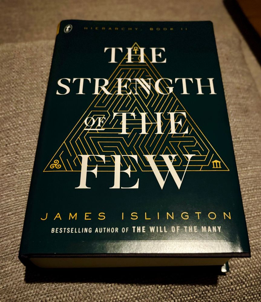 Hardback book titled The Strength of the Few by James Islington. The cover design features a geometric golden triangular maze pattern centered behind the title text. At the top, it notes that this is “Hierarchy: Book II”, indicating it’s the second book in a series. Along the bottom, it mentions that Islington is the bestselling author of The Will of the Many. The book has a dark green or teal cover with gold and white text.