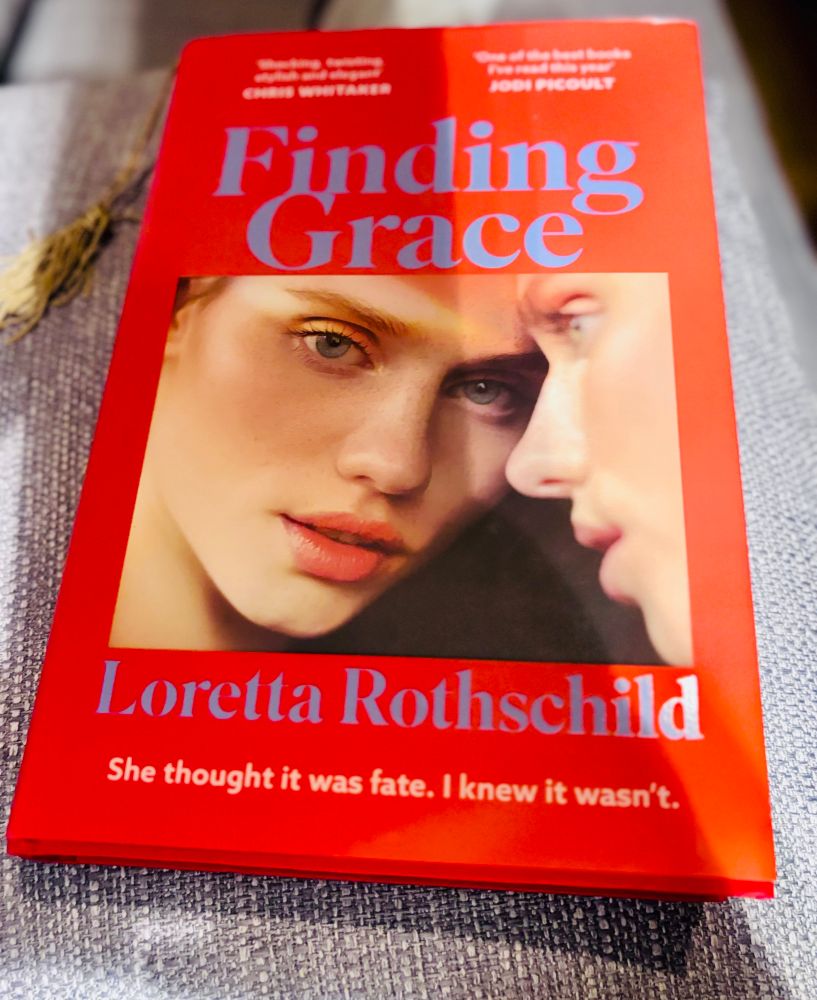 hardback book with a bright red cover.
book is "Finding Grace" in large, blue font.
The author's name is Loretta Rothschild, written in white text below the main image on the cover.
The cover also features a tagline: "She thought it was fate. I knew it wasn't." in white text.
There are also blurbs/quotes from other authors at the top:
 * One from Chris Whitaker mentioning "shocking, twisting, stylish and elegant."
 * Another from Jodi Picoult stating, "One of the best books I've read this year."
The cover image itself is a close-up of two faces, seemingly of women, in a somewhat intense or close proximity.
The book is resting on a textured surface that looks like a light-colored, woven fabric. There's also a bit of a dried, light-colored, straw-like decorative element visible on the left side of the image.

