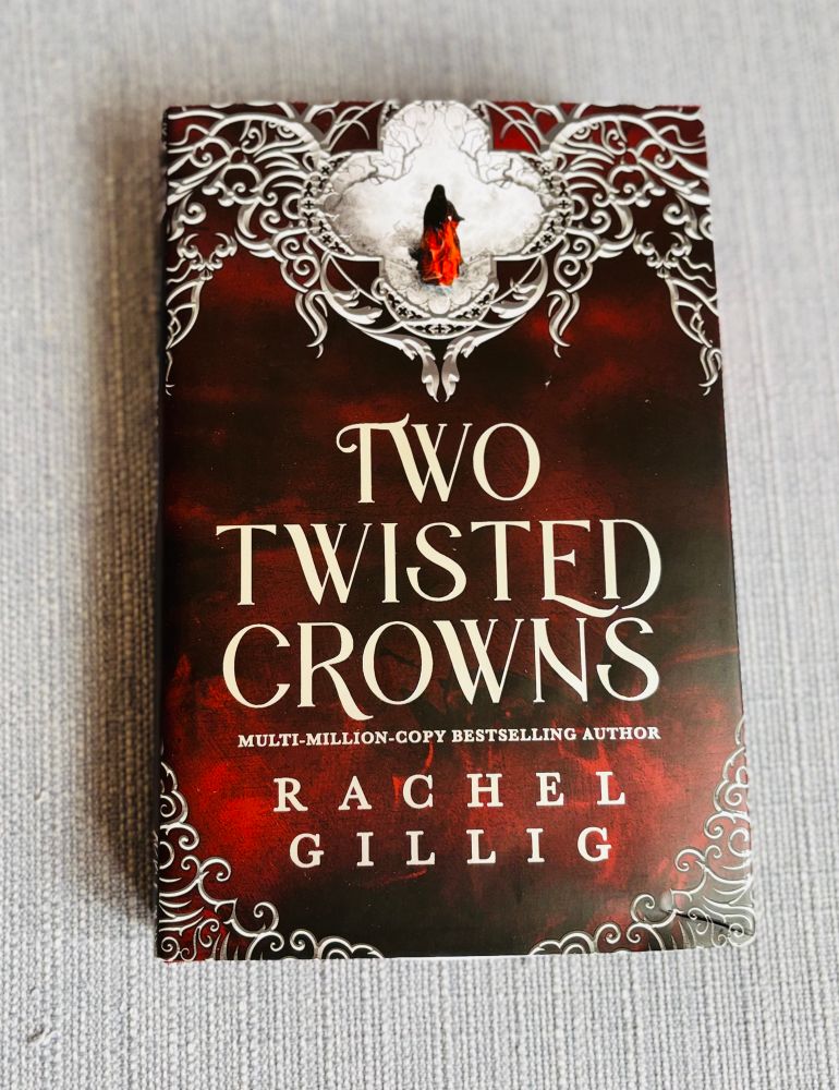 The photo shows the book Two Twisted Crowns by Rachel Gillig. The cover has a dark red and black color scheme with ornate, silver decorative patterns around the edges, similar to the design of One Dark Window. At the top, there’s an illustration of a figure in a flowing red cloak, framed within an intricate silver motif. The text on the cover reads:
	•	Title: Two Twisted Crowns
	•	Author: Rachel Gillig
	•	Tagline: Multi-million-copy bestselling author

This book is the sequel to One Dark Window.
