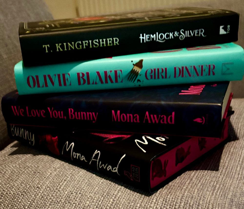 a stack of four books arranged on a grey fabric surface. From top to bottom, the titles and authors are:
	1.	“Hemlock & Silver” by T. Kingfisher
	2.	“Girl Dinner” by Olivie Blake
	3.	“We Love You, Bunny” by Mona Awad
	4.	“Bunny” by Mona Awad
