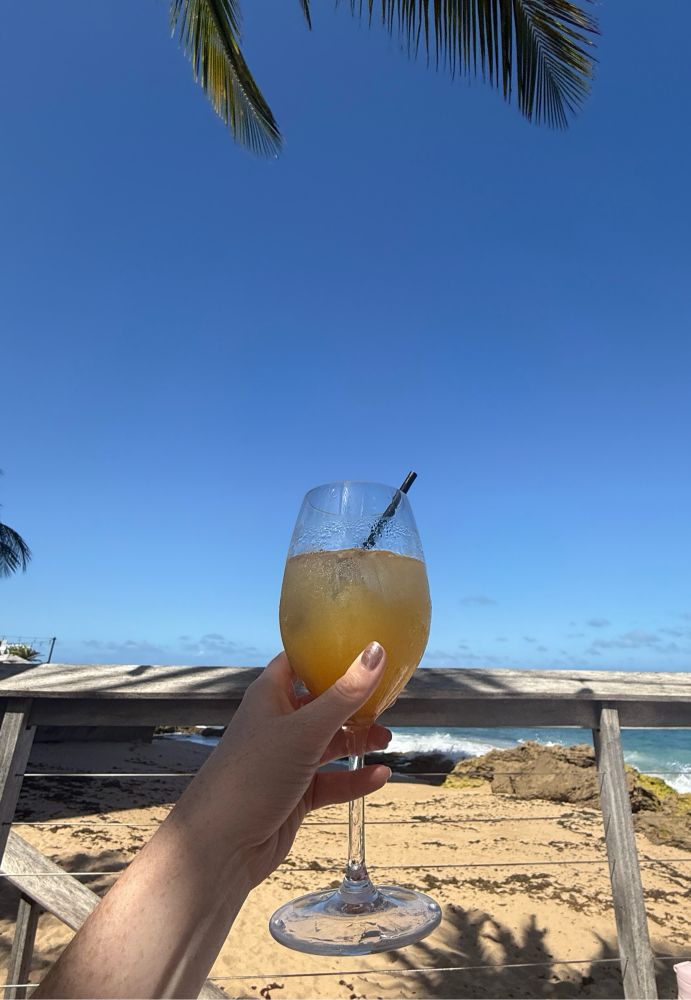 A hand lifting a goblet of white sangria with a beach and blue skies behind it. 