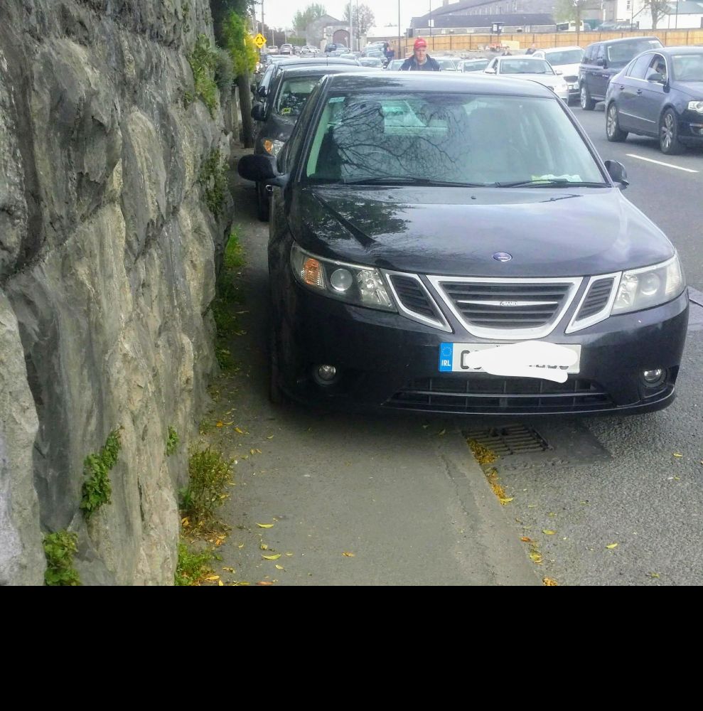 Car parked on a pavement with maybe 6 inches left for pedestrians to pass. Other similarly badly parked cars on both sides of a not very wide road.
