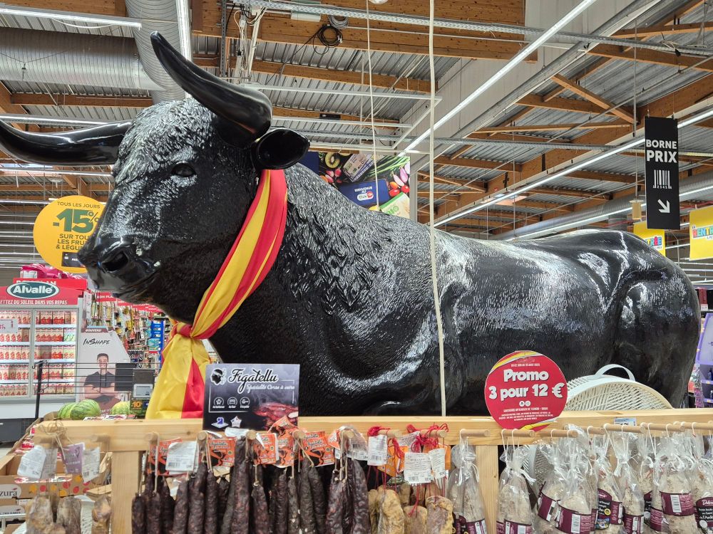 Photo taken in a French supermarket: a giant model of a bull advertises various bovine-pizzle sausages and testicle-cuts. A sign says "PROMO 3 POUR €12!"