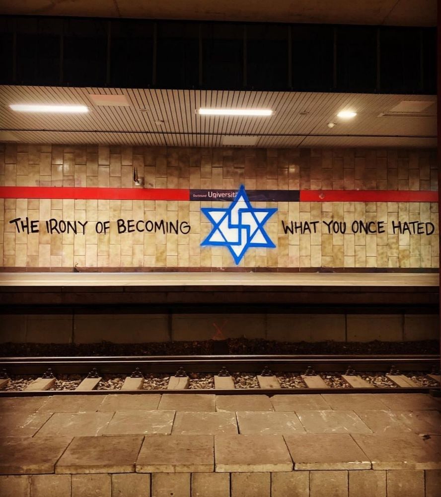 A subway with graffiti on the wall. A modified star of David encapsulated with the words "the irony of becoming * what you once hated"