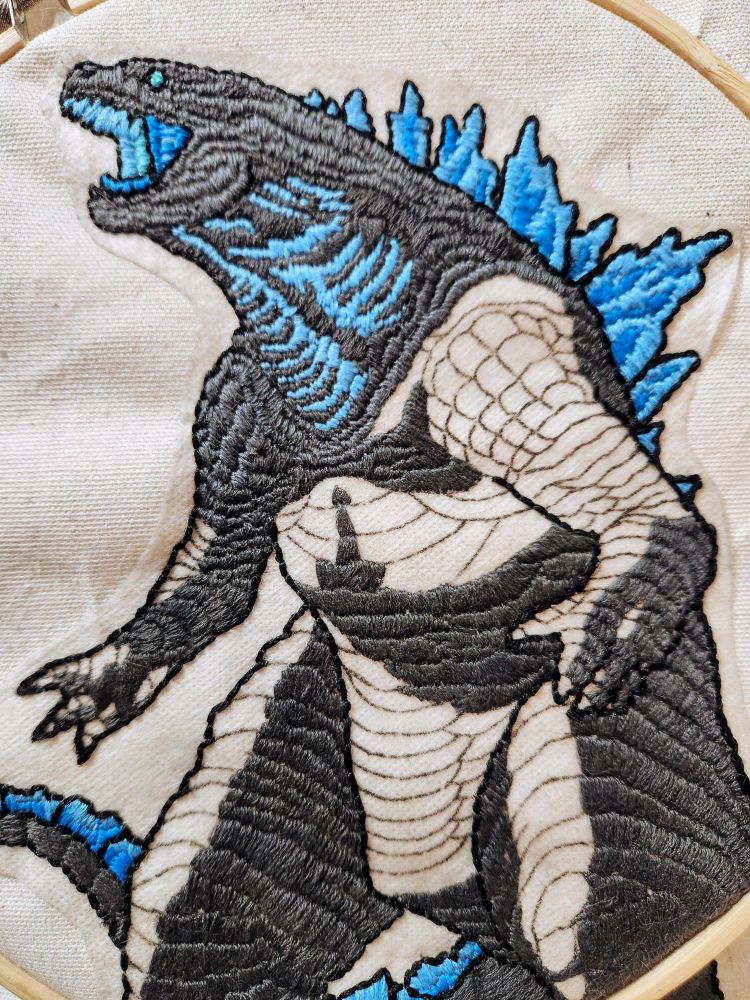 An image of an incomplete embroidered godzilla standing and roaring. His mouth and back spines are glowing blue and the completed portions of skin are represented as dark pewter-grey rolls.