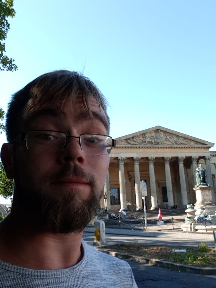 A surprised looking "Bobbi" in front of Victoria rooms in bristol wearing half rimmed glasses, a grey top. They have messy hair and a beard.