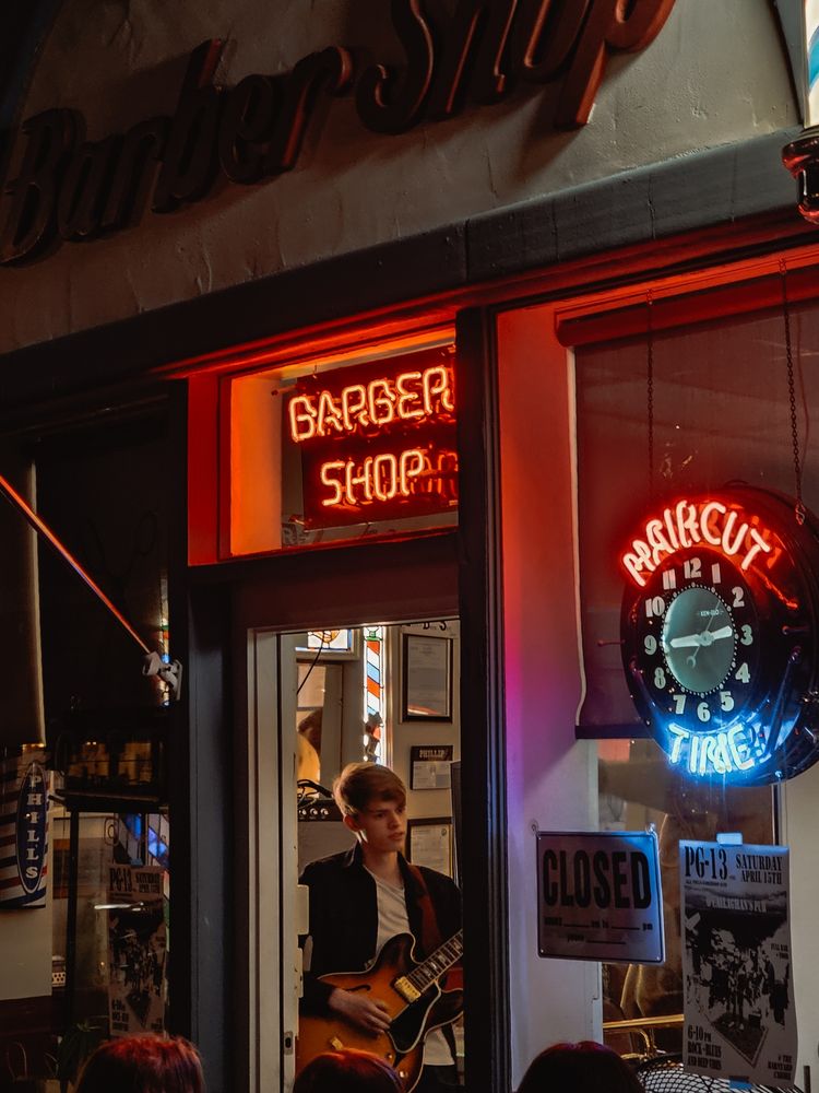 A live music performance taking place inside a barbershop, at night.
A band is performing, while the audience is seated on a bench outside, watching the performance through a window and open door.
Neon signs for "Barbershop" and "OPEN" are visible, along with a "PG-13" rating sign.
An American flag is partially visible in the foreground on the right.