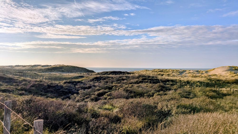 Foto van de duinen. Je ziet een klein stukje zee tussen de duinen door. De lucht is blauw met wat witte wolken 