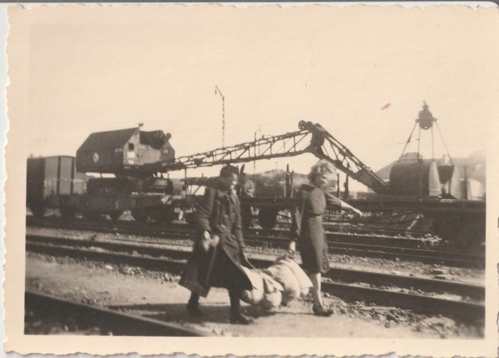 A man and woman carry a heavy bag in between them along rail lines. A huge gentry crane can be seen in the back. 