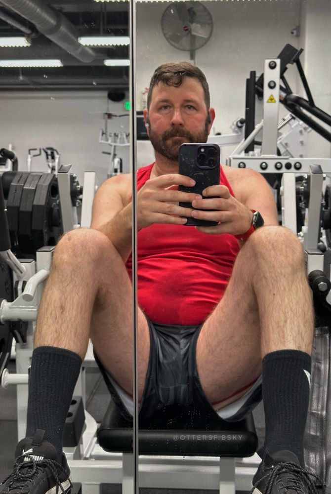 Me seated yet again on the hip thrust machine wearing a red tank top. Up the legs of my black camo shorts the red butt straps of my jock are visible.