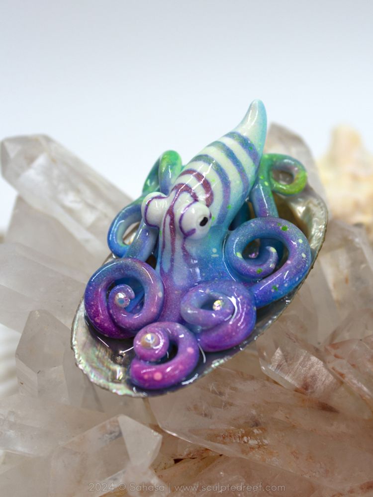 A colorful striped and speckled octopus sculpture, painted in blue, purple, hot pink and neon green. The octopus is tucked into a shell and holds tiny Swarovski crystals 