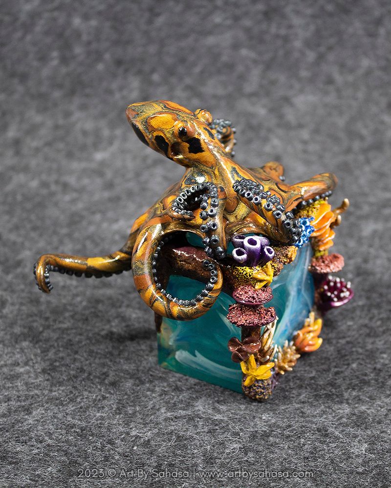 Photo of an octopus sculpture sitting on a miniature sculpted reef. The octopus is made from colored and effect clays that have been worked into a pattern that resembles the colors and banding of tiger's eye (the mineral). The plinth is a custom pour resin cube with transparent blue and teal green swirls with some white. Around the edges of the cube miniature corals and starfish have been added. They are handsculpted and painted.