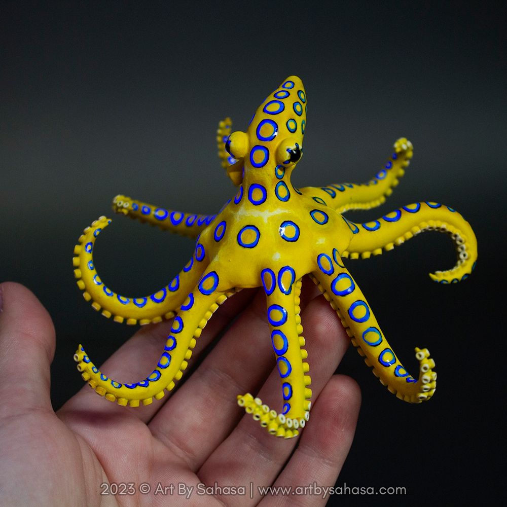 Photo of a blue ringed octopus sculpture sitting on the artist's hand. It's bright blue rings are activated by blacklight.