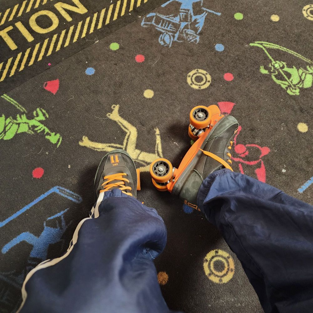 Lower legs of someone wearing black roller skates with orange laces and wheels. 
