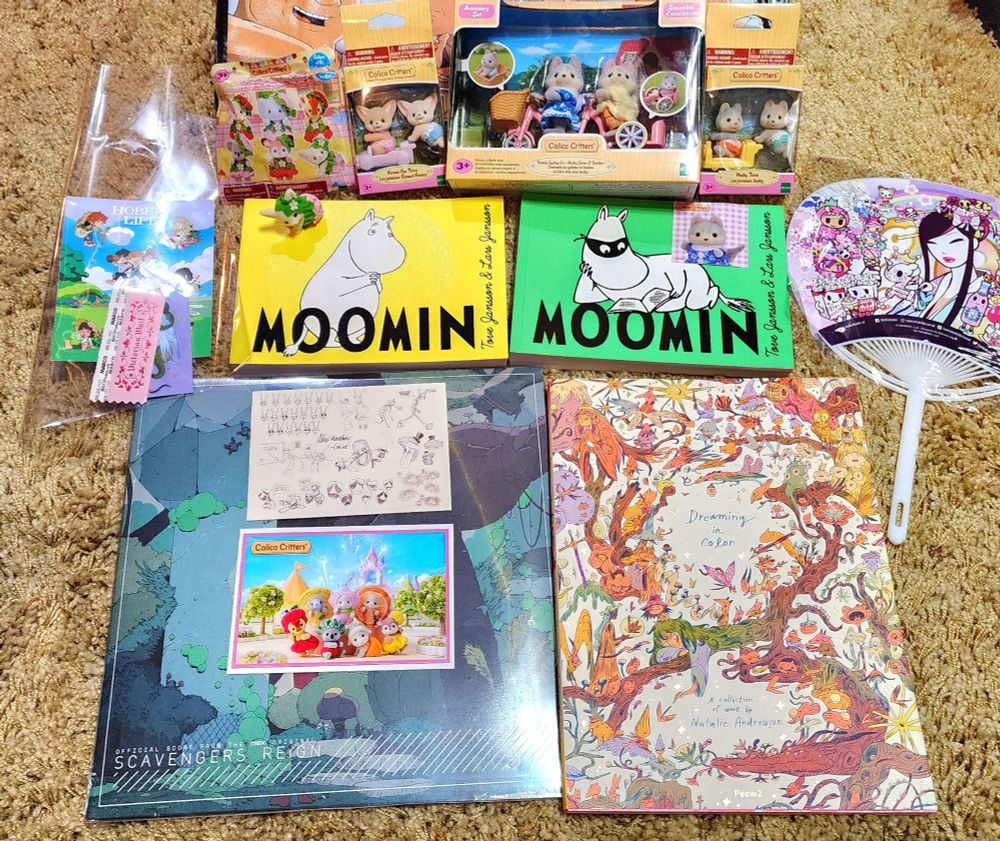 Photo of items bought from Comic Con, including a Scavengers Reign album, two Moomin volumes, an artbook, several Calico Critters, and an Amazing Digital Circus postcard filled with concept art signed by Alex Rochon (Caine)