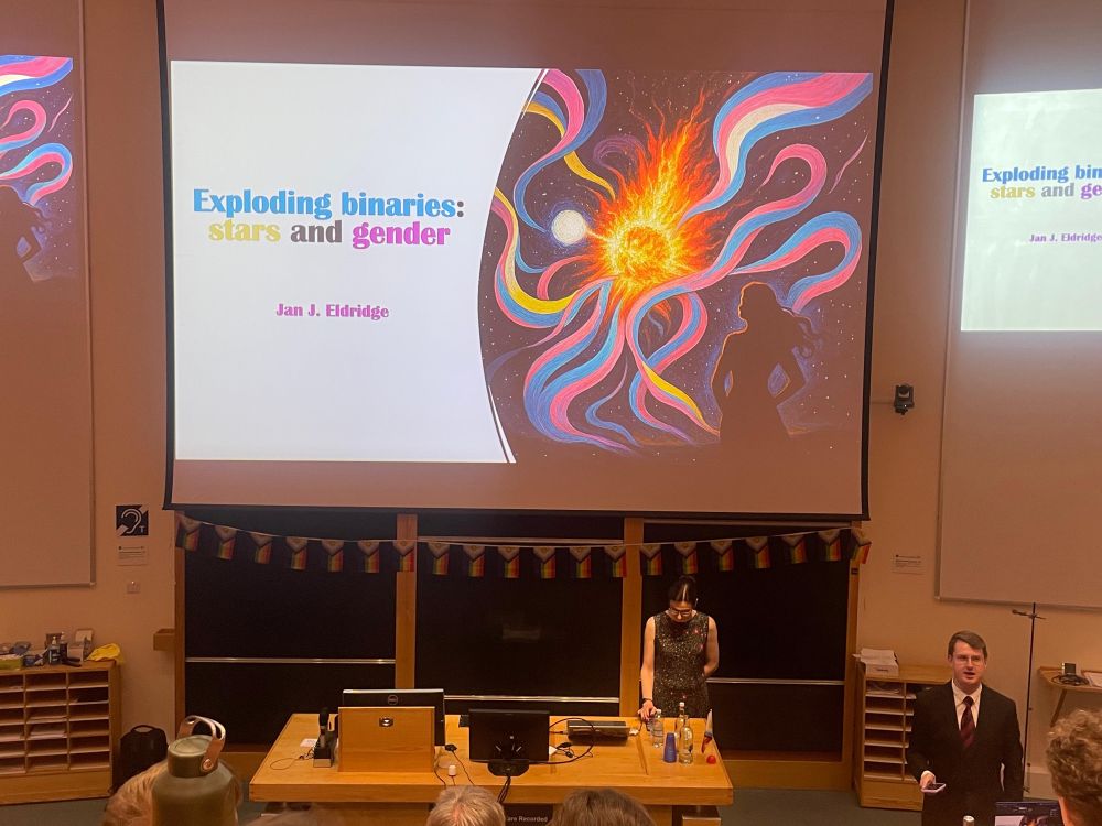Image taken in a lecture theatre, the screen displays the title of the talk “Exploding Binaries: stars and gender”, with a woman standing at the front of the lecture theatre 