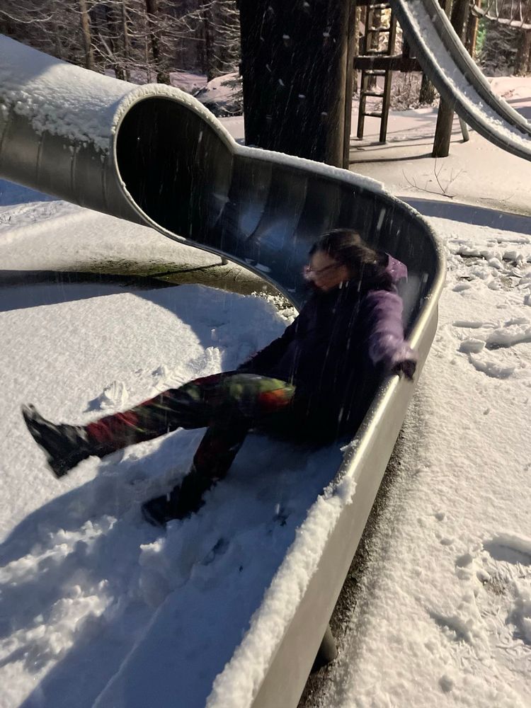 Snow covered playground with a metal slide. A dark haired woman in a purple jacket and flowery trousers is at the bottom of the slide laughing.