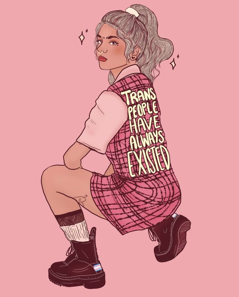 Woman with brown hair and blonde highlights, wearing a pink t-shirt and striped pink dress, heeled shoes with the trans flag on it. The dress has the words 'trans people have always existed' written on it. 