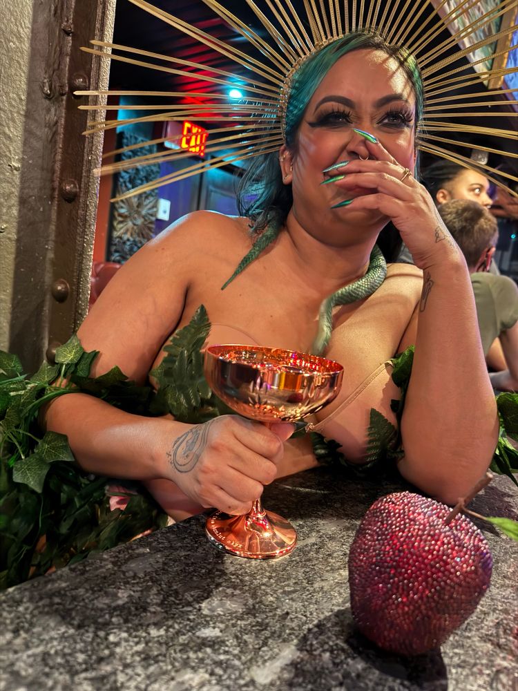 Dr. Bitch dressed in leaves as Eve in the garden sits at a bar holding a copper coupe glass. A crystal studded apple rests on the bar. Dr. Bitch is covering her mouth and giggling. 