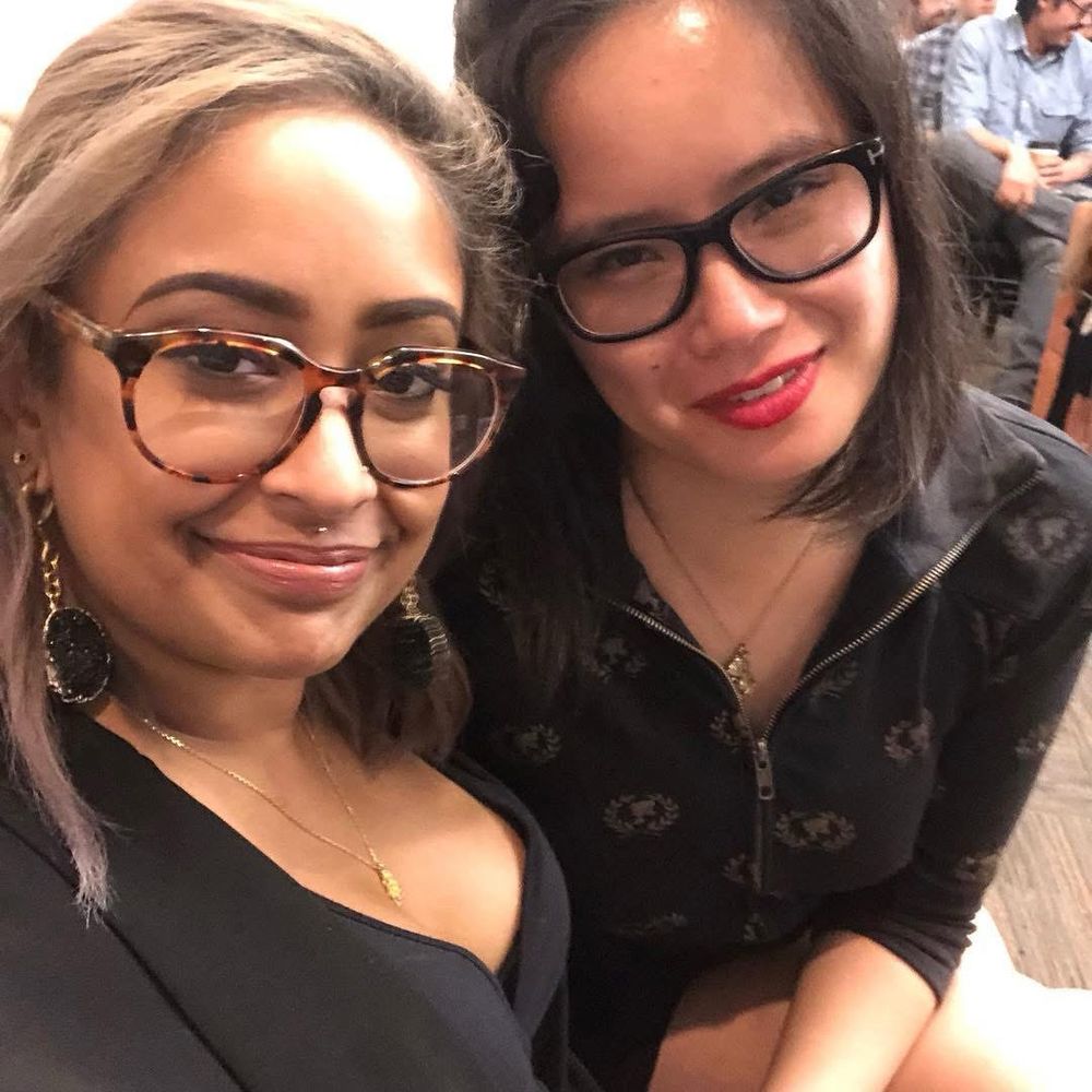 Rita Mookerjee and Dorothy K. Chan at AWP Tampa. Both femmes wear black tops and glasses. Chan wears red lipstick and a sparkly necklace. Mookerjee wears black druzy ear weights and a gold necklace. 