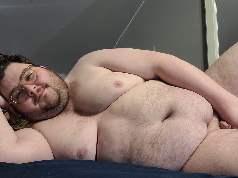 Chubby boy laying on his side posing naked, smiling slyly at the camera. Left arm over penis, right rested under head