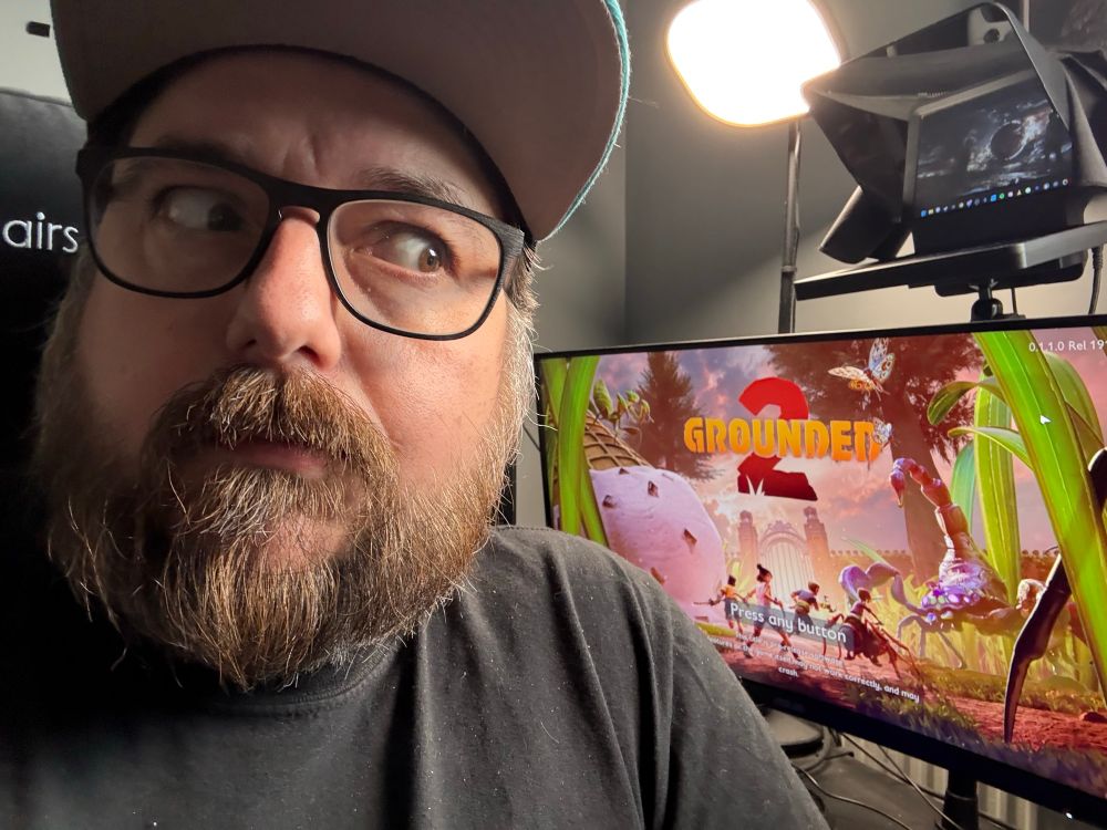 Close up shot of a man in a hat with a beard looking off to the side. Behind him is a computer monitor displaying the title menu for Grounded 2