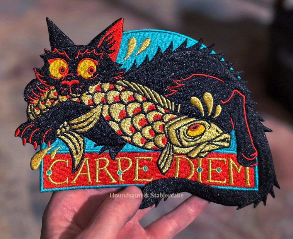 A large embroidered patch depicting a black cat catching a golden carp. The gold lettering at the bottom says “Carpe Diem”