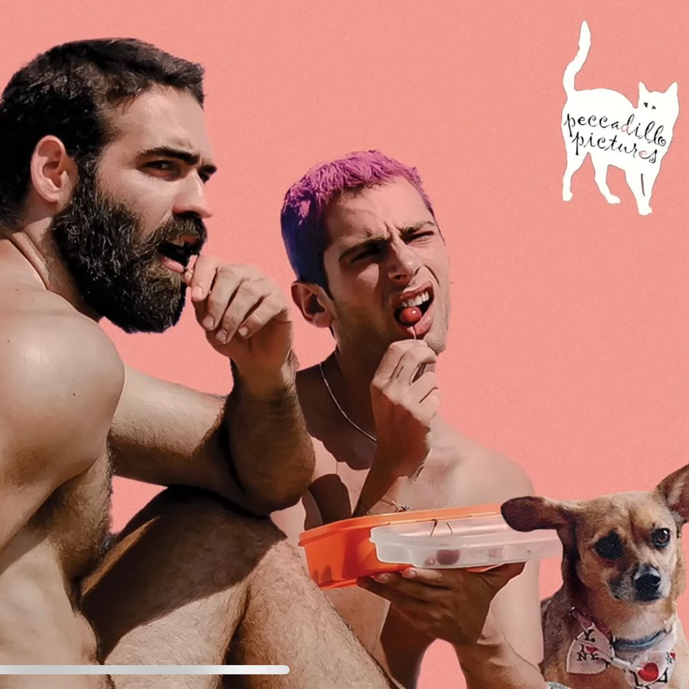 The image shows gay buddies Demosthenes and Nikitas, snacking on cherries as they lounge on a beach. Beside them, Carmen, a little mongrel, looks on soulfully. 