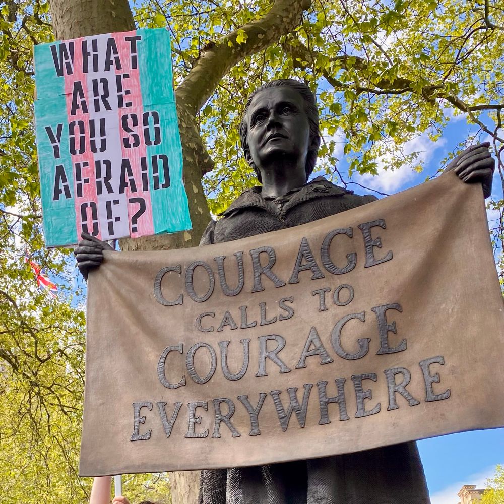 Statue of Suffragette leader Millicent Fawcett in Parliament Square. She holds a banner which reads: ‘Courage calls to courage everywhere.’ Behind her banner, a protestor’s arm reaches up to hold a pink-white-and-light-blue placard, which says: ‘What are you so afraid of?’