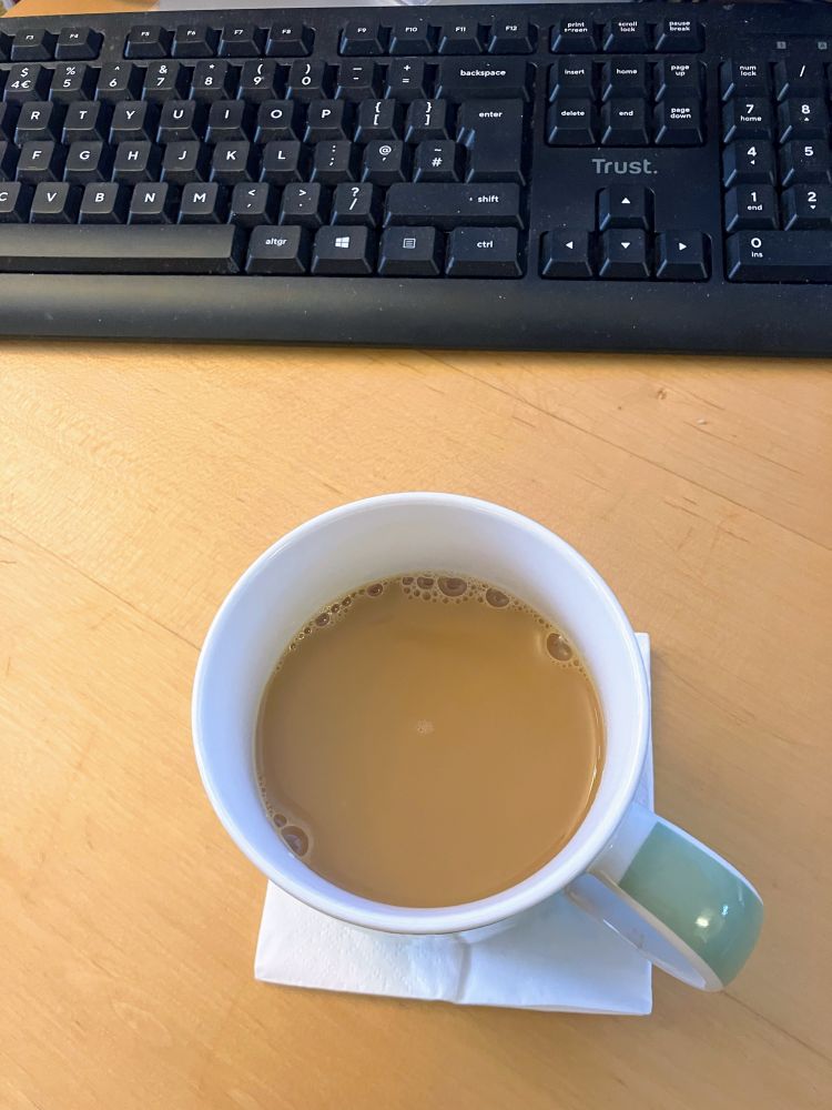 A tea cup on a paper tissue, near a keyboard, on a sandy colored desk.