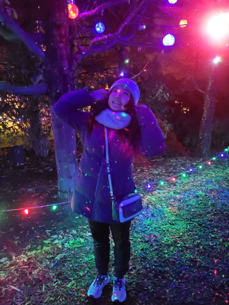A photo of me, tucking hair behind my ears looking up at Christmas lights underneath a tree, sparkles of lights on my jacket